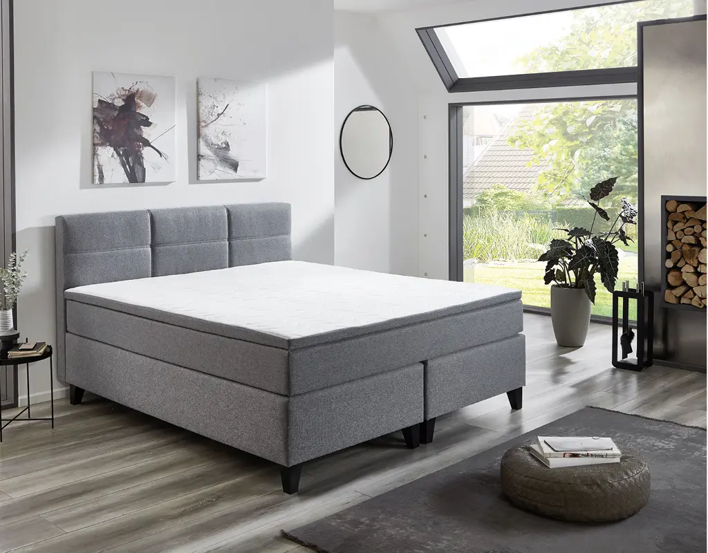 Modernes graues Boxspringbett in hellem Schlafzimmer mit großen Fenstern, Wandbildern und stilvollen Pflanzen. Komfortables Bett für erholsamen Schlaf und moderne Einrichtung.